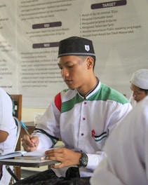 A young man wearing a traditional cap and a white shirt with green and red accents is writing in a notebook. He appears to be in a classroom or study setting, with a poster featuring text in the background.