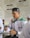 A young man wearing a traditional cap and a white shirt with green and red accents is writing in a notebook. He appears to be in a classroom or study setting, with a poster featuring text in the background.