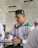 A young man wearing a traditional cap and a white shirt with green and red accents is writing in a notebook. He appears to be in a classroom or study setting, with a poster featuring text in the background.