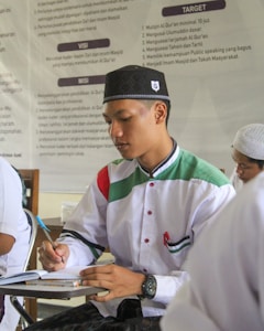 A young man wearing a traditional cap and a white shirt with green and red accents is writing in a notebook. He appears to be in a classroom or study setting, with a poster featuring text in the background.
