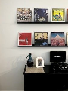 Shelves display various vinyl records with album art featuring diverse designs. Below is a black table with a vintage-style record player, a dark ceramic pitcher with a unique handle, and a small framed picture.