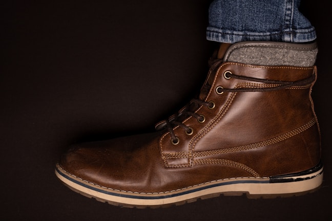 A brown leather boot with visible stitching details, eyelets for laces, and a textured sole. The boot is worn with a pair of blue jeans, partially visible at the top.