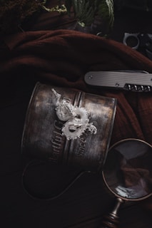 A vintage metal mug with embossed text sits on a dark wooden surface. On top of it lies a detailed, silver dragon ornament. Nearby, a closed pocket knife with a metallic grip and a magnifying glass rest, partially draped by a brown fabric. Faint foliage is visible in the background, adding an earthy feel.