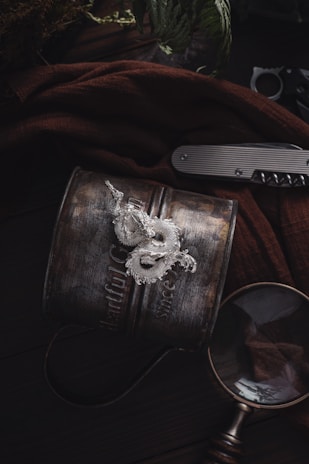 A vintage metal mug with embossed text sits on a dark wooden surface. On top of it lies a detailed, silver dragon ornament. Nearby, a closed pocket knife with a metallic grip and a magnifying glass rest, partially draped by a brown fabric. Faint foliage is visible in the background, adding an earthy feel.