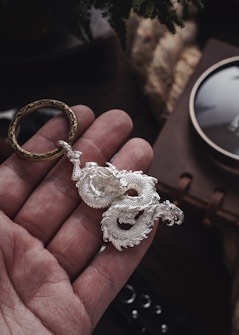 A detailed silver dragon pendant rests in the palm of a hand, held by a brass ring with intricate designs. The pendant features textured scales and a sinuous shape, while the background is subtly blurred with hints of a leather-bound book and soft lighting effects.