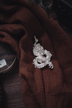 A silver dragon-shaped ornament rests on a textured brown fabric, which is draped elegantly. The background includes dark wood surfaces and a circular object partially visible, suggesting an antique or vintage theme.