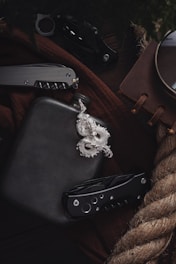 A collection of items including a black flask, a silver dragon pendant, two folding knives, a brown notebook, and a piece of thick rope, arranged on a dark brown fabric surface.