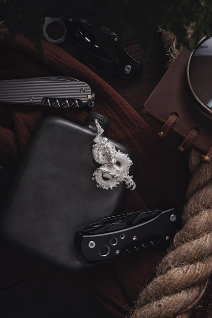 A collection of items including a black flask, a silver dragon pendant, two folding knives, a brown notebook, and a piece of thick rope, arranged on a dark brown fabric surface.