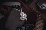 A silver dragon pendant lies on a dark surface, accompanied by a black flask. Nearby, a folding knife with a textured handle is partially visible. A brown notebook with leather binding and a large magnifying glass are placed on a coarse rope. The items suggest a theme of adventure or exploration.