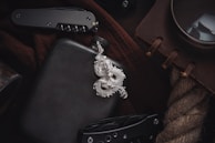 A silver dragon pendant lies on a dark surface, accompanied by a black flask. Nearby, a folding knife with a textured handle is partially visible. A brown notebook with leather binding and a large magnifying glass are placed on a coarse rope. The items suggest a theme of adventure or exploration.