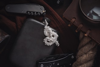 A silver dragon pendant lies on a dark surface, accompanied by a black flask. Nearby, a folding knife with a textured handle is partially visible. A brown notebook with leather binding and a large magnifying glass are placed on a coarse rope. The items suggest a theme of adventure or exploration.