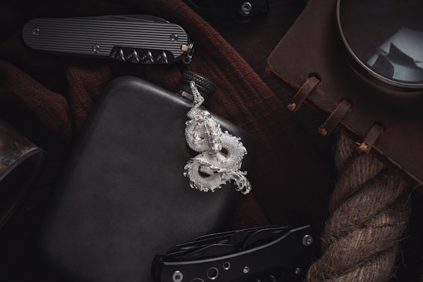 A silver dragon pendant lies on a dark surface, accompanied by a black flask. Nearby, a folding knife with a textured handle is partially visible. A brown notebook with leather binding and a large magnifying glass are placed on a coarse rope. The items suggest a theme of adventure or exploration.
