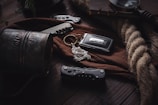 An arrangement of items on a wooden table includes a dark metallic mug with engraved text, pocket knives, a car key with a Jaguar logo, a coil of rope, and a brown fabric creating an intricate composition.