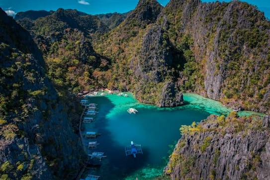 A breathtaking aerial view of a turquoise lagoon surrounded by steep, verdant cliffs. Several traditional boats are floating on the calm water, adding a sense of tranquility and adventure to the scene. The vibrant greenery contrasts sharply with the deep blue and green hues of the water.