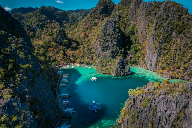 A breathtaking aerial view of a turquoise lagoon surrounded by steep, verdant cliffs. Several traditional boats are floating on the calm water, adding a sense of tranquility and adventure to the scene. The vibrant greenery contrasts sharply with the deep blue and green hues of the water.