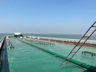 A large cargo ship's deck with a wide, green-painted metal surface extending into the distance. Several structural elements and machinery are visible along the deck, with white railings on both sides. The horizon shows a vast body of water under a clear blue sky, and a distant land or structure is visible.