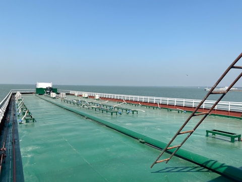 A large cargo ship's deck with a wide, green-painted metal surface extending into the distance. Several structural elements and machinery are visible along the deck, with white railings on both sides. The horizon shows a vast body of water under a clear blue sky, and a distant land or structure is visible.