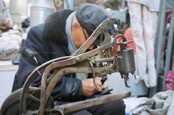 An elderly person, wearing a cap and a coat with a fur collar, is closely examining or working with a vintage sewing machine. Surrounding the individual are a mixture of fabrics and other sewing materials, suggesting a setting related to tailoring or garment repair.
