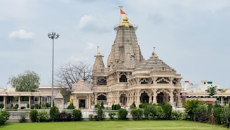 A detailed view of an intricately designed Hindu temple with multiple spires, ornate carvings, and a flag atop the tallest spire. The temple is surrounded by lush greenery and well-maintained gardens. In the foreground, there are small hedges and a grassy area, while the sky above is partially cloudy.