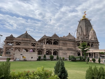 A grand and intricately designed temple with detailed carvings adorns the landscape, featuring multiple domed structures and a large, centrally prominent shikhar. The temple's ornate architecture exhibits a traditional style, set against a backdrop of a partly cloudy blue sky. In the foreground, a well-maintained garden with green grass and various shrubs enhances the scene.