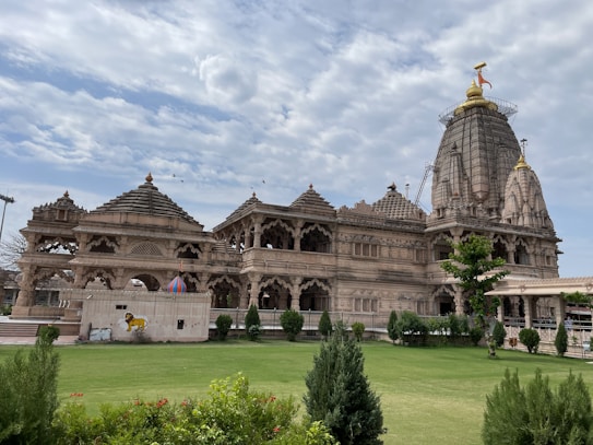 A grand and intricately designed temple with detailed carvings adorns the landscape, featuring multiple domed structures and a large, centrally prominent shikhar. The temple's ornate architecture exhibits a traditional style, set against a backdrop of a partly cloudy blue sky. In the foreground, a well-maintained garden with green grass and various shrubs enhances the scene.