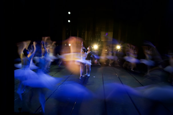 Ballet dancers on stage are captured in motion, creating a mesmerizing blend of blurred movements and light trails against a dimly lit background. The scene exudes a dynamic energy with spotlights highlighting the center dancers.