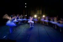 A group of ballet dancers in motion on a dimly lit stage, with two central figures in focus wearing traditional ballet attire. The surrounding dancers appear blurred, creating a dynamic sense of movement and energy. Bright stage lights illuminate the scene from the background, casting reflections on the floor.