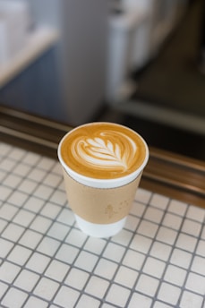 A cup of coffee with intricate latte art is placed on a tiled surface. The coffee cup has a cardboard sleeve for insulation and is positioned in a warm, inviting environment.