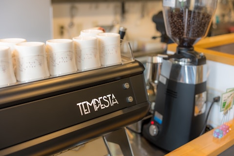 A coffee counter displays a sleek espresso machine branded 'Tempesta' alongside several white ceramic cups. To the right, a coffee grinder filled with beans is visible, and the setting is a well-organized cafe environment.