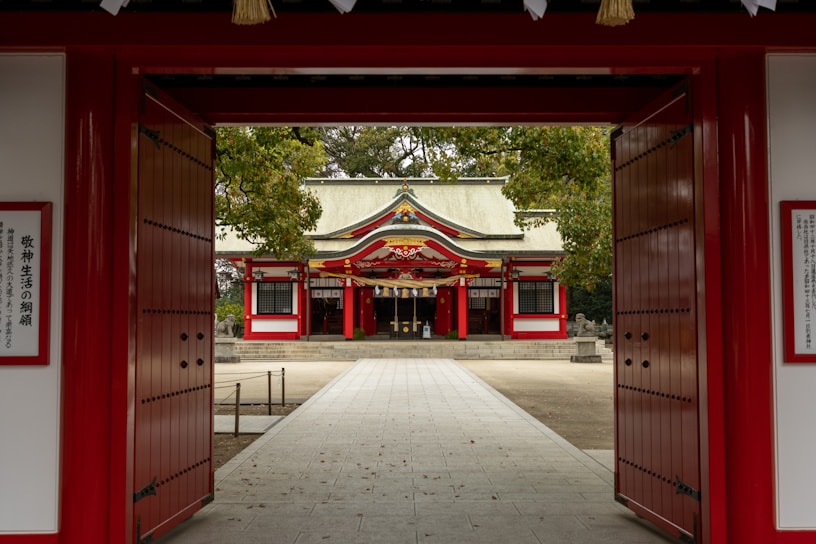 A traditional Japanese shrine is visible through large, open red gates. The structure has a distinctive roof with ornate decorations and is surrounded by lush greenery. The pathway leading to the shrine is paved and maintained, contributing to the serene and peaceful ambiance of the setting.