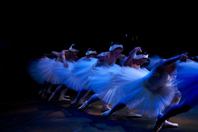 A group of ballet dancers performing a synchronized routine on stage. The dancers are dressed in white tutus and headpieces, and the stage is dark with low lighting, creating a dramatic effect. The dancers are in a line, tilting forward with one arm extended, creating a sense of motion and grace.