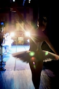 A ballerina in a tutu stands backstage, silhouetted against bright stage lights. Other dancers are visible in the background, preparing for a performance. The scene captures the anticipation and focus of a dance production.