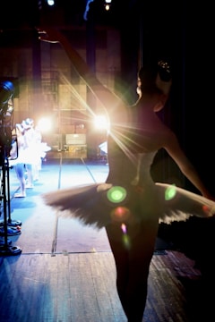 A ballerina in a tutu stands backstage, silhouetted against bright stage lights. Other dancers are visible in the background, preparing for a performance. The scene captures the anticipation and focus of a dance production.