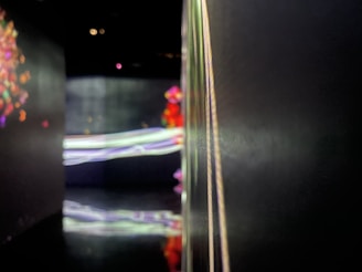 A dimly lit, abstract scene with blurred lines of fluorescent colors creating a dreamy atmosphere. Elements of light and shadows play on a reflective surface, suggesting a modern artistic installation.