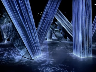 Geometric structures with illuminated surfaces create a futuristic environment. The lighting is primarily blue, casting intricate patterns and reflections across the floor and surrounding surfaces.