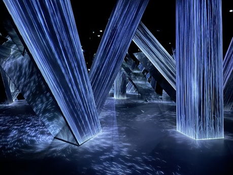 Geometric structures with illuminated surfaces create a futuristic environment. The lighting is primarily blue, casting intricate patterns and reflections across the floor and surrounding surfaces.