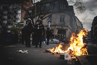 A street scene with debris burning intensely in the foreground. Three individuals in protective gear, possibly police officers or riot control, stand near the flames. The background displays an urban environment with buildings, bare trees, and onlookers.