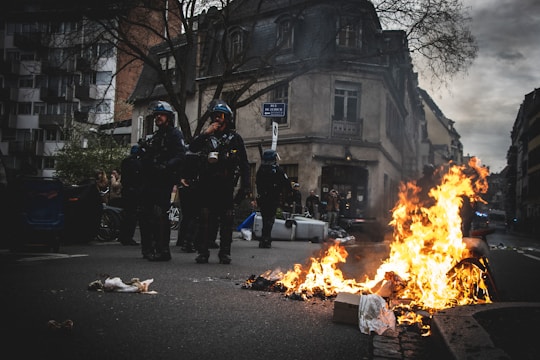A street scene with debris burning intensely in the foreground. Three individuals in protective gear, possibly police officers or riot control, stand near the flames. The background displays an urban environment with buildings, bare trees, and onlookers.
