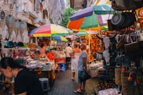 A vibrant market scene with colorful umbrellas providing shade over stalls filled with various goods. Dreamcatchers, leather products, and decorative bull skulls are prominently displayed. People are browsing items, adding to the bustling atmosphere with a mix of locals and tourists.