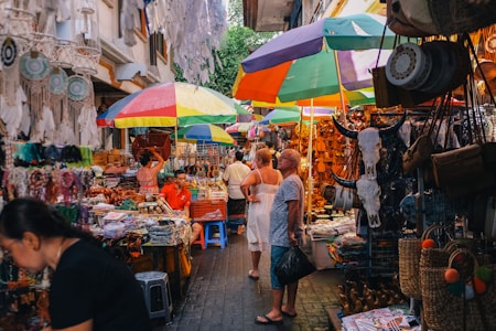 A vibrant market scene with colorful umbrellas providing shade over stalls filled with various goods. Dreamcatchers, leather products, and decorative bull skulls are prominently displayed. People are browsing items, adding to the bustling atmosphere with a mix of locals and tourists.