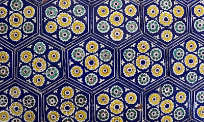 Hexagonal tiles featuring intricate floral patterns in vibrant colors are arranged in a repeating geometric pattern. Each tile contains stylized flowers with yellow, green, and white petals surrounding a central circular design, set against a deep blue background.