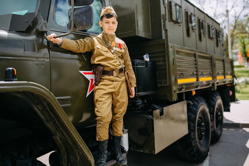 A young boy in a military-style uniform stands beside a large military truck. The uniform includes a hat and several medals on the chest. The truck is dark green with a red star emblem on the side.
