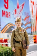 A young boy dressed in a military uniform stands in front of a backdrop featuring a star and Cyrillic letters. He wears several medals and carries a brown satchel.