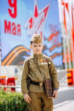 A young boy dressed in a military uniform stands in front of a backdrop featuring a star and Cyrillic letters. He wears several medals and carries a brown satchel.