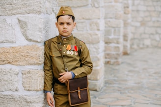 A young boy dressed in a military-style uniform stands against a stone wall. He wears several medals and a small red star on his cap. A brown satchel hangs from his shoulder, and his expression is serious.
