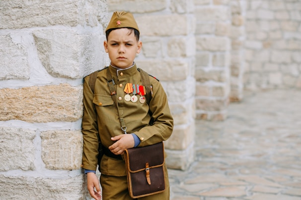 A young boy dressed in a military-style uniform stands against a stone wall. He wears several medals and a small red star on his cap. A brown satchel hangs from his shoulder, and his expression is serious.