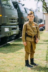 A young boy wearing a vintage military uniform stands on grassy ground with military trucks lined up behind him. He has several medals pinned to his chest and is holding a notebook or booklet. There is a tree and a building visible in the background.