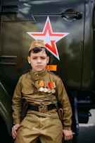 A boy dressed in a military-style uniform adorned with various medals stands beside a vehicle marked with a red star emblem. His expression is serious, and his attire includes a cap that matches his khaki-colored outfit. The image conveys a theme related to military or historical commemoration.
