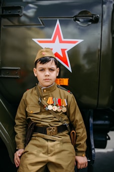 A boy dressed in a military-style uniform adorned with various medals stands beside a vehicle marked with a red star emblem. His expression is serious, and his attire includes a cap that matches his khaki-colored outfit. The image conveys a theme related to military or historical commemoration.