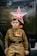 A boy dressed in a military-style uniform adorned with various medals stands beside a vehicle marked with a red star emblem. His expression is serious, and his attire includes a cap that matches his khaki-colored outfit. The image conveys a theme related to military or historical commemoration.
